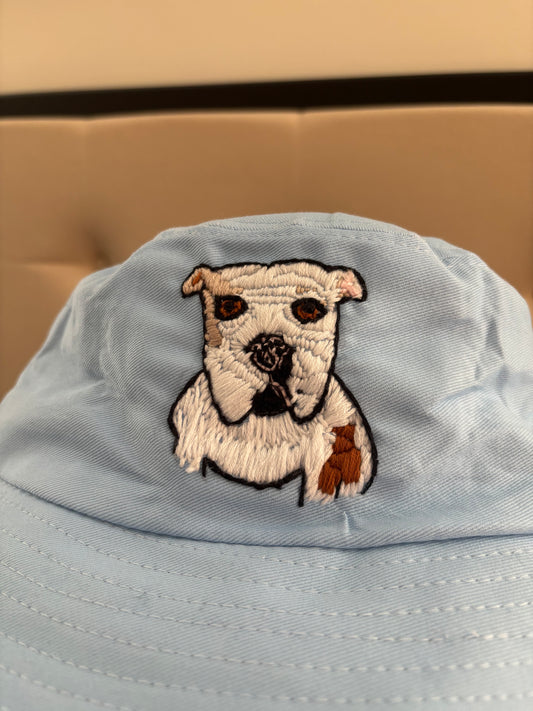 Portrait of white dog with brown eyes and brown and tan spots on a blue bucket hat in front of a beige background